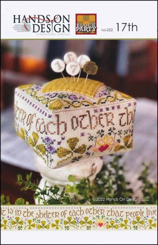 Cube-shaped St. Patrick’s Day–themed cross-stitch pincushion with floral motifs, green and gold accents, and the proverb “It is in the shelter of each other that people live,” displayed on a pedestal with decorative pins.