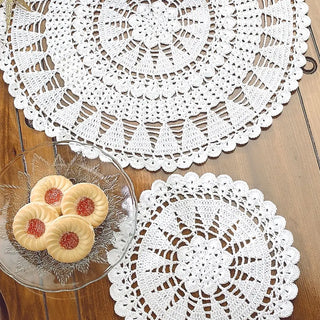 Two white crocheted doilies on a wooden surface with a glass plate of cookies.