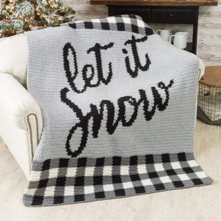 Gray crochet blanket with 'let it snow' text on a white couch, Christmas tree in the background