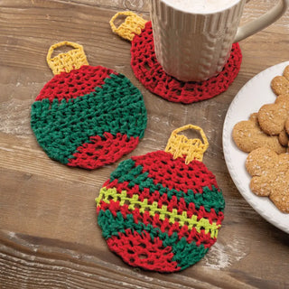 Two crocheted Christmas ornament coasters on a wooden surface with a mug and cookies.