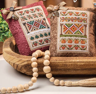 Two cross-stitched pillows with autumn patterns in a wooden bowl, accompanied by a beaded necklace.