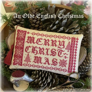 Red monochromatic cross-stitch design reading “Merry Christmas,” bordered with traditional sampler motifs, small trees, a star, and decorative banding, finished as a rectangular pillow displayed with pinecones and Christmas décor.