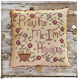A small cross-stitched pillow with the phrase “Pray For Me I’m Anxious,” surrounded by rose and plum flowers on green vines, with a small stitched basket in the lower left. Finished with scalloped trim and displayed on rustic wood with scattered buttons.
