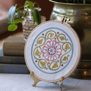Embroidery hoop with floral design on a stand, books, and plant in the background