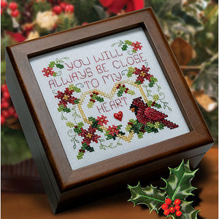 Framed cross-stitched artwork with decorative text and holly leaves on a wooden surface.