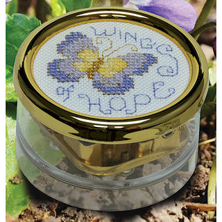 Cross-stitched butterfly design in a clear container with gold lid, surrounded by greenery.