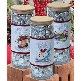 Three glass jars with wooden lids filled with silver candies, featuring winter-themed labels.