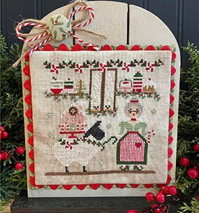 A cross stitch design of a festive farmhouse scene featuring a woman in a red dress holding a peppermint stick beside a white sheep on a holly-trimmed leash, surrounded by Christmas decorations and candles.