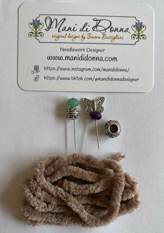A needlework design set by Mani di Donna, featuring three decorative pins (green bead, butterfly, purple bead, and a round bezel), a coiled bundle of tan chenille stem cord, and a branded white card.