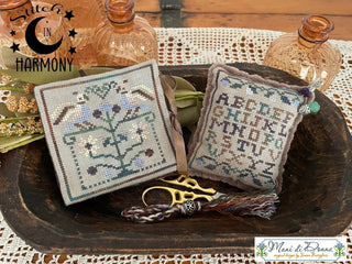 Two finished cross-stitch sampler pillows displayed on a rustic wooden tray with scissors and decorative glass bottles. The left sampler features a tree with birds and flowers, and the right sampler shows an alphabet patchwork design.