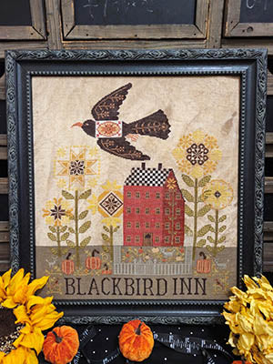 Blackbird Inn Cross Stitch Pattern by Quaint Rose Needle Arts – stitched design featuring a black-and-white bird in flight above sunflowers with “Blackbird Inn” lettering on rustic light fabric.