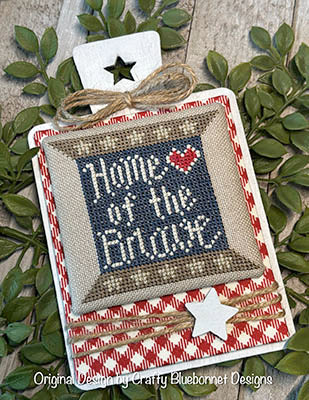 A patriotic cross stitch design reading “Home of the Brave” in white lettering on a gray square with a stitched red heart above, framed in warm tan tones for a rustic Americana look.