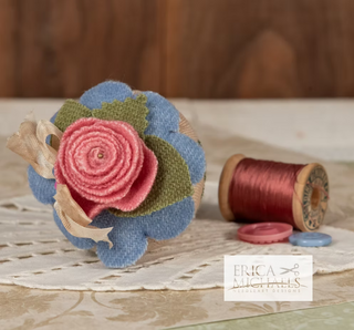 Decorative pincushion with pink flower and blue background, next to a spool of red thread on a wooden surface.