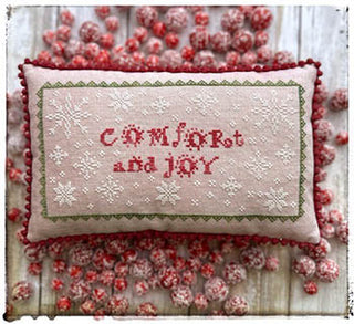 A small rectangular cross-stitched pillow on blush-pink fabric with the words “Comfort and Joy” surrounded by white snowflakes and framed by a green stitched border. Finished with red pom trim and displayed atop sugared red berry decorations.