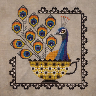 Fancy cross stitch pattern by The Artsy Housewife featuring a stylized peacock with colorful fanned tail feathers sitting in a patterned bowl, framed by a decorative border.