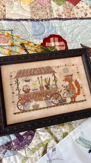 Framed cross-stitched piece of a fox with a flower cart on a quilted background