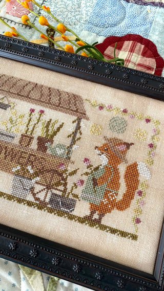 Cross-stitched scene with a fox and garden elements in a decorative frame.