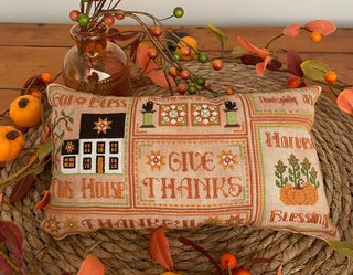 Rectangular autumn-themed cross stitch pillow featuring a white colonial-style house, pumpkins, harvest motifs, and decorative lettering including "God Bless This House," "Give Thanks," and "Harvest Blessings" stitched in warm fall colors.