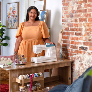 Woman in an orange dress standing next to a sewing machine on a wooden table with a brick wall background.