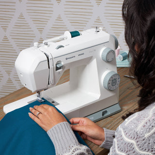 Person using a white sewing machine on a wooden table with a patterned wall in the background.