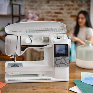White sewing machine on a wooden table with a blurred background of a woman and decorative items.