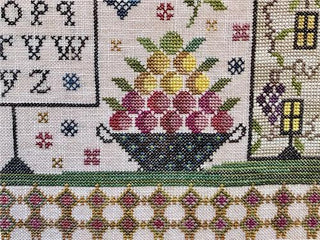 Cross-stitched design with a bowl of fruit and flowers, surrounded by alphabets and numbers.