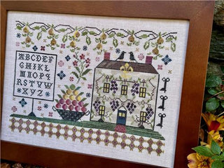 Decorative cross-stitched panel with a house, alphabet chart, and floral elements in a wooden frame.