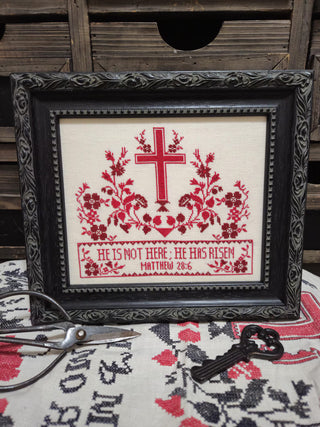 Framed cross-stitched embroidery with red floral design and text, placed on a wooden surface.