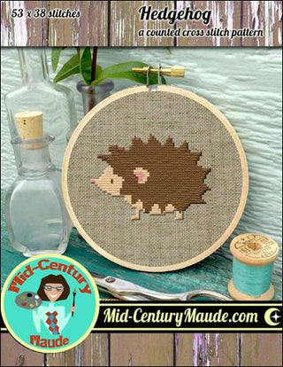 Cross stitch pattern of a hedgehog with materials and tools on a wooden surface, featuring Mid-Century Maude branding.