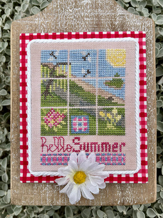 A decorative cross-stitch piece mounted on a rustic wooden plaque, featuring a 3x3 summer scene with a windmill, house, flowers, and the words 'Hello Summer' in pink embroidery, accented with a white daisy flower in front.