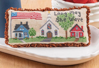 Decorative pillow with cross-stitched design of houses and an American flag, placed on a white surface.