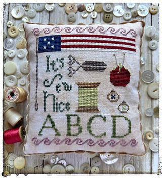 A small cross-stitched pillow with a patriotic American flag, sewing motifs (needle, pincushion, spool, scissors), the phrase “It’s Sew Nice,” and the letters A B C D, displayed on a wooden surface surrounded by buttons and thread spools.