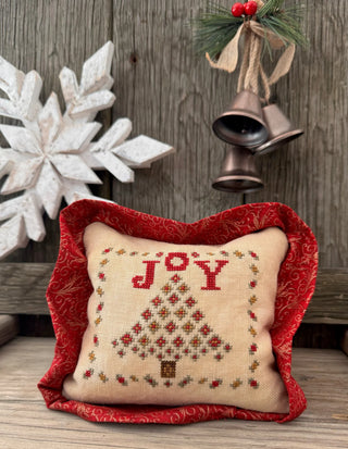 Decorative pillow with 'JOY' and Christmas tree design, surrounded by festive decorations on a wooden background.