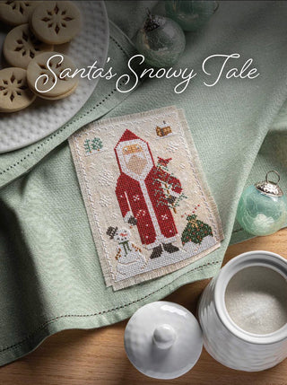 A needlepoint canvas featuring Santa Claus in a snowy scene, displayed on a sage green cloth with holiday-themed props including wooden ornaments, glass ornaments, and ceramic containers. The text 'Santa's Snowy Tale' is written across the top.