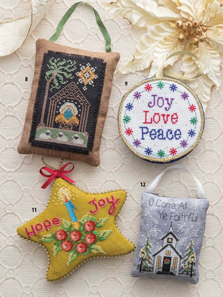 Four decorative cross-stitch ornaments displayed on a cream textured fabric background with dried flowers. The ornaments include a rectangular brown pouch with a green hanging ribbon, a round white ornament reading 'Joy Love Peace,' a yellow star-shaped ornament with 'Hope Joy' and a candle motif, and a gray hanging ornament with a church illustration and the text 'O Come All Ye Faithful.
