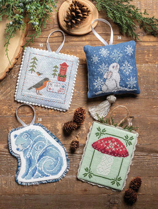 Four handmade embroidered Christmas ornaments displayed on a rustic wooden surface with pinecones and evergreen branches. The ornaments include a square patch with a lighthouse and bird, a square patch with a rabbit and snowflakes, a curved patch with a blue wave pattern, and a square patch with a red and white mushroom.