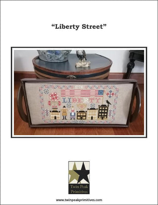 Cross-stitched pillow with 'Liberty Street' design on a wooden surface, featuring Twin Peak Primitives branding.