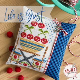 Cross-stitch design featuring jars filled with red berries and a sampler-style layout with colorful patterns, finished as a decorative pillow with coordinating fabric.
