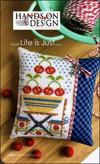 Cross-stitch design featuring jars filled with red berries and a sampler-style layout with colorful patterns, finished as a decorative pillow with coordinating fabric.