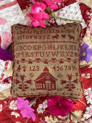 Little pinkeep pillow with a sampler cross stitch pattern stitched in red on vintage colored fabric with a little red schoolhouse in the center at the bottom with the date 1862 stitched on either side of the schoolhouse.