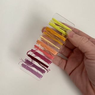 Hand holding a clear plastic card with colorful embroidery threads against a white background