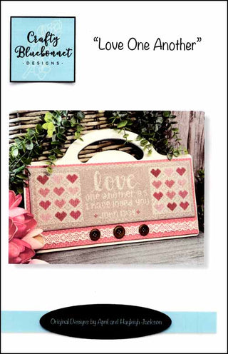 Cross stitch pattern featuring the phrase “Love One Another” with surrounding heart motifs in soft red and neutral tones, finished as a decorative pillow by Crafty Bluebonnet Designs.