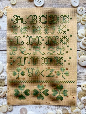 A small alphabet sampler stitched in green thread on tan 14ct perforated paper, featuring A–Z letters, scattered decorative motifs, and three shamrocks at the bottom, displayed on a rustic wood surface with white buttons.