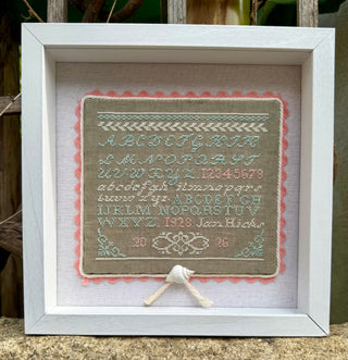 A framed vintage-style cross-stitch sampler featuring an alphabet, numbers, and decorative borders. The sampler is mounted on a beige fabric background with a pink scalloped edge, set inside a white wooden frame.