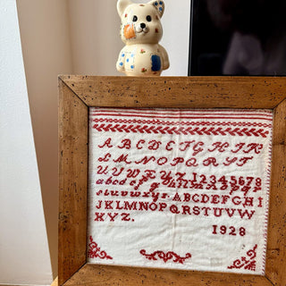 A framed vintage-style embroidered amigurumi needlework sampler with red lettering on white fabric, enclosed in a rustic wooden frame. A small cream-colored teddy bear figurine sits on top of the frame against a light wall.