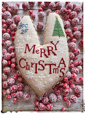 A tall primitive heart-shaped stitched pillow with “Merry Christmas,” white snowflakes, a small Christmas tree, and “Dec 25th,” displayed on a rustic wood surface surrounded by sugared red berries.