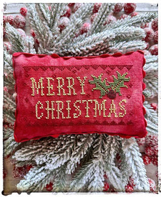 Small red cross-stitched pillow with the words ‘Merry Christmas’ and a holly sprig, designed by Lucy Beam, displayed on frosted greenery.