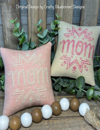 Two decorative pillows with 'Mom' text on a wooden surface with greenery