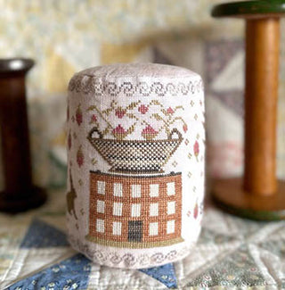 Cylindrical cross-stitched drum featuring a brick-red house, floral vine motifs, and a decorative strawberry in a bowl on top, photographed on a quilt backdrop with wooden spools.