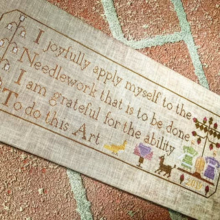 An inspirational cross stitch sampler featuring a quote about joyful needlework surrounded by delicate floral and folk-art motifs in soft muted colors, shown in two different colorways.